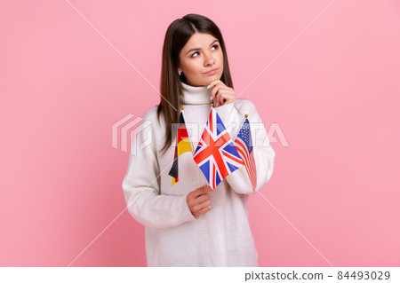 Thoughtful young girl holds flags in hands, looks away with pensive expression, studying languages, wearing white casual style sweater. Indoor studio shot isolated on pink background. Thoughtful young girl holds flags in hands, looks away with pensive expression, studying languages, wearing white casual style sweater. Indoor studio shot isolated on pink background. 84493029