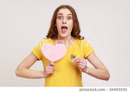 Portrait of young surprised woman in yellow casual T-shirt holding pink heart on stick and physical bitcoin, has shocked face expression, cryptocurrency. Indoor studio shot isolated on gray background 84493030