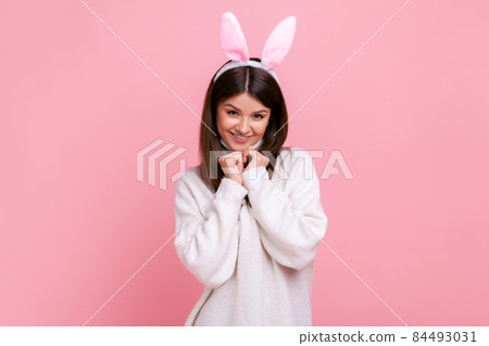 Portrait of cute charming young adult brunette woman with bunny ears, looking at camera with smile, wearing white casual style sweater. Indoor studio shot isolated on pink background. Portrait of cute charming young adult brunette woman with bunny ears, looking at camera with smile, wearing white casual style sweater. Indoor studio shot isolated on pink background. 84493031