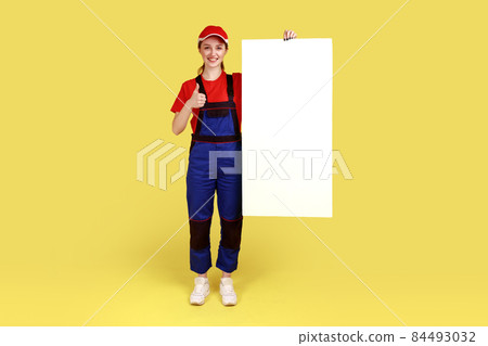 Full length portrait of worker woman standing with blank poster with copy space for your advertisement, showing thumb up, wearing overalls and red cap. Indoor studio shot isolated on yellow background Full length portrait of worker woman standing with blank poster with copy space for your advertisement, showing thumb up, wearing overalls and red cap. Indoor studio shot isolated on yellow background 84493032