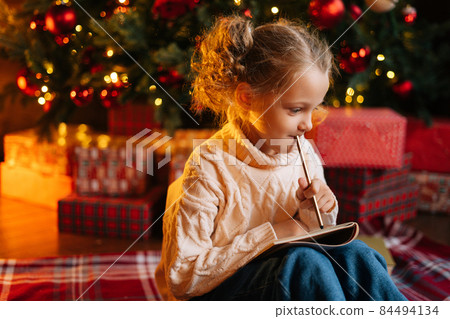 Side view of happy thoughtful little blonde girl writing letter to Santa Claus sitting on floor on background of Christmas tree and gift boxes Side view of happy thoughtful little blonde girl writing letter to Santa Claus sitting on floor on background of Christmas tree and gift boxes 84494134