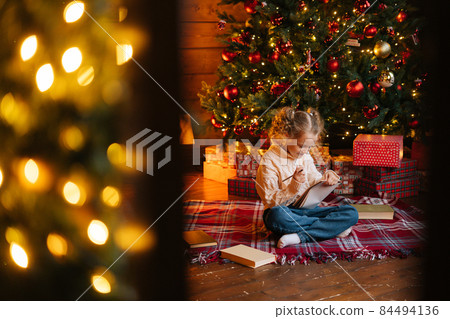 Remote view of smiling charming little blonde curly child girl writing letter to Santa Claus sitting on floor sitting by Christmas tree and gift boxes 84494136