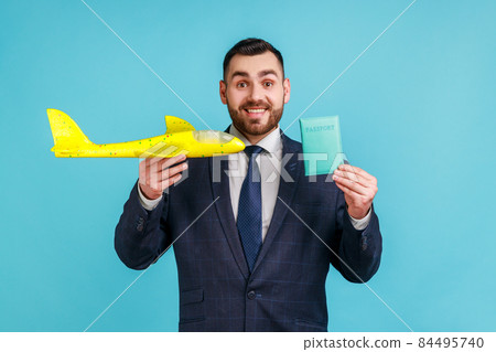 Positive man wearing official style suit holding passport and paper airplane, looking at camera with happy expression, satisfied by journey. Indoor studio shot isolated on blue background. Positive man wearing official style suit holding passport and paper airplane, looking at camera with happy expression, satisfied by journey. Indoor studio shot isolated on blue background. 84495740