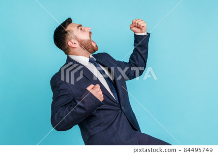 Side view of overjoyed handsome man wearing official style suit expressing winning gesture with raised fists and screaming, celebrating victory. Indoor studio shot isolated on blue background. Side view of overjoyed handsome man wearing official style suit expressing winning gesture with raised fists and screaming, celebrating victory. Indoor studio shot isolated on blue background. 84495749