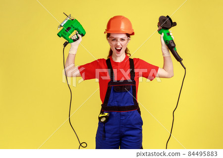 Excited crazy builder woman standing with fretsaw and grinding saw in hands and screaming loudly, wearing work uniform and protective helmet. Indoor studio shot isolated on yellow background. Excited crazy builder woman standing with fretsaw and grinding saw in hands and screaming loudly, wearing work uniform and protective helmet. Indoor studio shot isolated on yellow background. 84495883