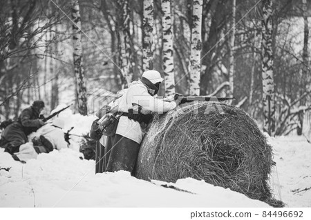 Re-enactor Dressed Like German Wehrmacht Infantry Soldier In World War II Aiming Rifle from Hay roll At Winter Day. Historical Re-enactment. Black And White Photo. WWII 84496692