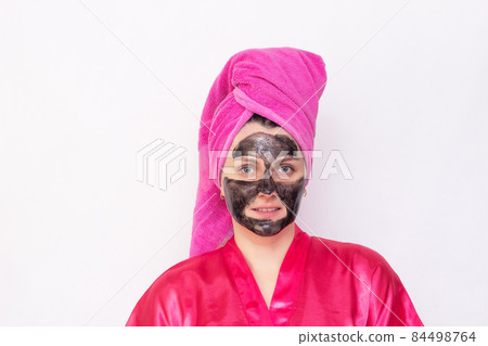 A girl on a white background with a black mask on her face against blackheads and black dots in a pink robe and a towel on her head, copy space A girl on a white background with a black mask on her face against blackheads and black dots in a pink robe and a towel on her head, copy space 84498764
