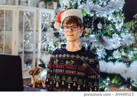 Portrait of Millenial, gen Z young man in glasses and Santa Claus hat sitting with laptop and celebrating Christmas online with friends on the Christmas tree background. Virtual party. Video chat Portrait of Millenial, gen Z young man in glasses and Santa Claus hat sitting with laptop and celebrating Christmas online with friends on the Christmas tree background. Virtual party. Video chat 84499523
