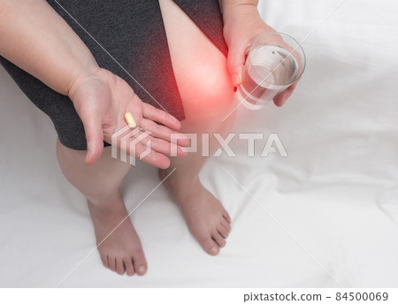 A woman with a sore knee holds a chondroprotector tablet and a glass of water, glucosamine and chondroitin sulfate in her hand, copy space A woman with a sore knee holds a chondroprotector tablet and a glass of water, glucosamine and chondroitin sulfate in her hand, copy space 84500069