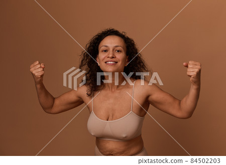 Delighted smiling dark-haired curly woman in beige underwear, clenching her fists, smiling with a toothy smile, looking at the camera. Body positivity, concept of combating oppression of women rights Delighted smiling dark-haired curly woman in beige underwear, clenching her fists, smiling with a toothy smile, looking at the camera. Body positivity, concept of combating oppression of women rights 84502203