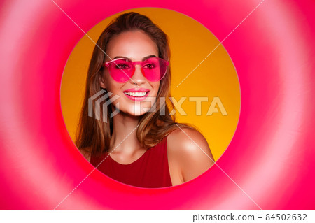 Portrait of cheerful young woman in pink sunglasses who holds inflatable ring over yellow background Portrait of cheerful young woman in pink sunglasses who holds inflatable ring over yellow background 84502632