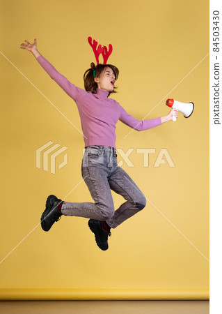 Full-length portrait of excited young woman cheerfully shouting in megaphone and jumping isolated over yellow background. Winter season 84503430