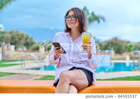 Middle-aged woman relaxing near pool using headphones and smartphone 84503537