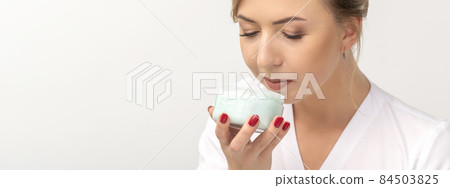 Portrait of young beautician sniffing moisturizing cream holding white jar on white background, copy space. Portrait of young beautician sniffing moisturizing cream holding white jar on white background, copy space. 84503825