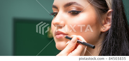 Makeup professional artist or cosmetologist is painting contour lips of a young woman with a pencil close up. 84503835