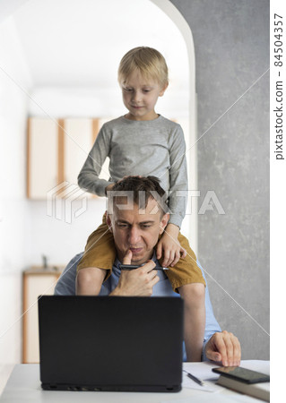Son sits on his dads shoulders while he works at the laptop. Young father tries to work at home with the children Son sits on his dads shoulders while he works at the laptop. Young father tries to work at home with the children 84504357