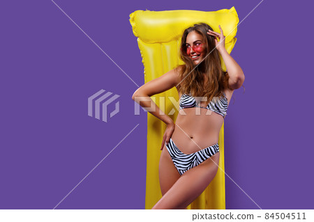 young woman posing in bikini with yellow inflatable mattress while standing on purple background young woman posing in bikini with yellow inflatable mattress while standing on purple background 84504511