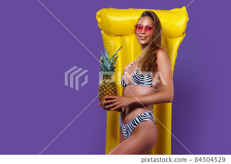 young slim caucasian girl posing in bikini with yellow inflatable mattress while holding pineapple.  84504529