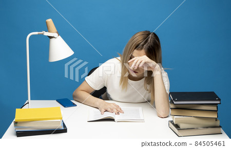 Tired young student woman in white casual clothes sitting at the table reading the book in library of university or college. Sitting and reading on blue background. Studying. 84505461