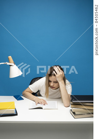 Tired young student woman in white casual clothes sitting at the table reading the book in library of university or college. Sitting and reading on blue background. Studying. 84505462