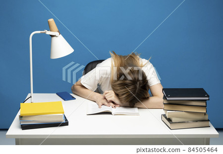 Tired student girl with glasses sleeping on the books in the library. Student studying hard exam and sleeping on books on a blue background. 84505464