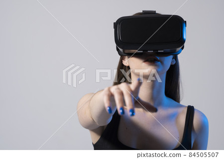 Brunette woman in a black t-shirt and VR headset looking up and trying to touch objects in virtual reality. VR is a technology that simulates a physical presence and to interact with environment. Brunette woman in a black t-shirt and VR headset looking up and trying to touch objects in virtual reality. VR is a technology that simulates a physical presence and to interact with environment. 84505507