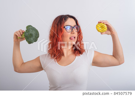 Caucasian woman chooses between vegetables and fast food. Redhead girl holding broccoli and donut on a white background. Caucasian woman chooses between vegetables and fast food. Redhead girl holding broccoli and donut on a white background. 84507579
