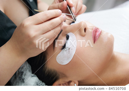 Eyelash extension procedure with tweezers in beauty salon Eyelash extension procedure with tweezers in beauty salon 84510708