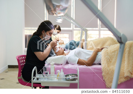 Professionals working with a clients in beaty salon. Woman lying on cosmetologiacal coach during lashes enlarging 84511030