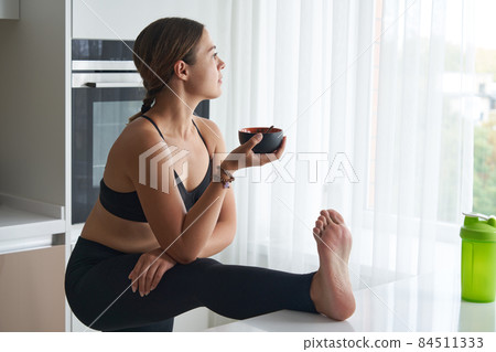 Young woman eating while stretching in kitchen with leg on table Young woman eating while stretching in kitchen with leg on table 84511333