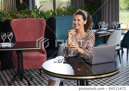 Beautiful young curly woman sitting in a cafe drinking fresh orange juice and waiting for her friend Beautiful young curly woman sitting in a cafe drinking fresh orange juice and waiting for her friend 84511472