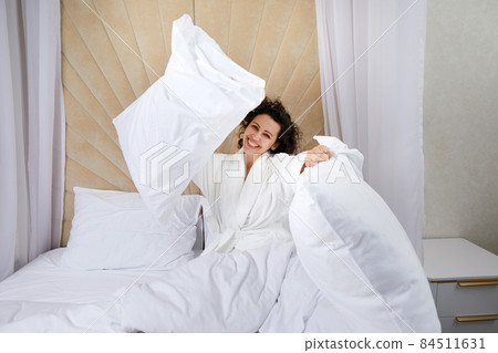 Beautiful young woman sits on the bed and throws a pillow in hotel having fun on weekend at home Beautiful young woman sits on the bed and throws a pillow in hotel having fun on weekend at home 84511631