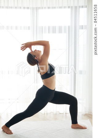 Portrait of beautiful young woman working out at home, doing yoga or pilates exercise. Standing in Warrior one pose, Virabhadrasana Portrait of beautiful young woman working out at home, doing yoga or pilates exercise. Standing in Warrior one pose, Virabhadrasana 84511658