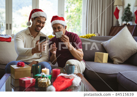 Happy biracial adult son and senior father in santa hats making smartphone christmas video call 84515639
