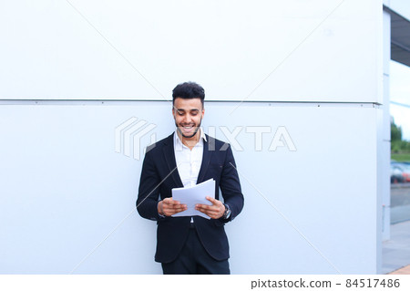 Arabian handsome student smiling, holds and looks at documents near wall of business center. Arabian handsome student smiling, holds and looks at documents near wall of business center. 84517486