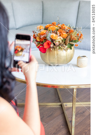 Brunette woman is taking a photo of a cute floral bouquet inside a cabbage-painted pumpkin. 84518085