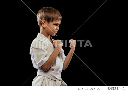 Cropped side view portrait of little boy in white kimono standing with fists up isolated over black background Cropped side view portrait of little boy in white kimono standing with fists up isolated over black background 84521441