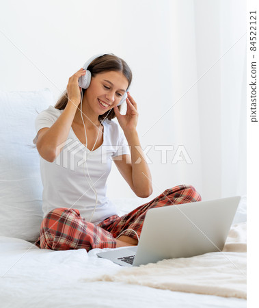 Young smiling woman sitting on bed in bedroom and listening to music or watching movie 84522121