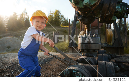 Cute cheerful child looking at camera and smiling while tightening bolt on pipe in oil field. Joyful kid in construction helmet using industrial wrench while repairing pump jack pipe on sunny day. 84523121