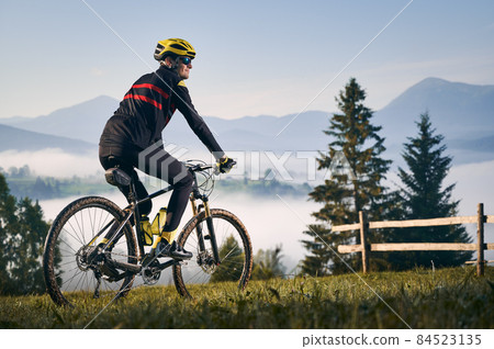 Back view of young man in cycling suit riding bicycle on grassy hill and looking at beautiful misty mountains. Male bicyclist in safety helmet enjoying the view of mountains during bicycle ride. 84523135