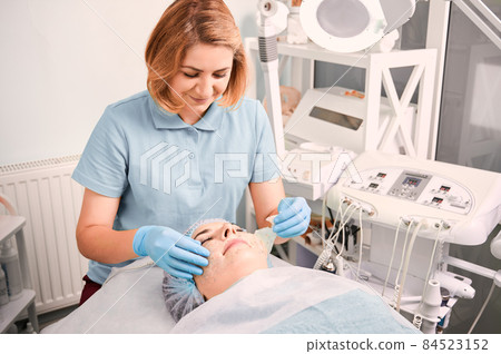 Cosmetologist removing deep cleansing mask from female client face. Young woman lying on daybed while having skincare procedure at wellness medical center. Concept of beauty and facial treatment. Cosmetologist removing deep cleansing mask from female client face. Young woman lying on daybed while having skincare procedure at wellness medical center. Concept of beauty and facial treatment. 84523152