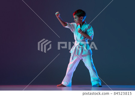 Full-length portrait of sportive child, training martial arts isolated over gradient blue background in neon Full-length portrait of sportive child, training martial arts isolated over gradient blue background in neon 84523815