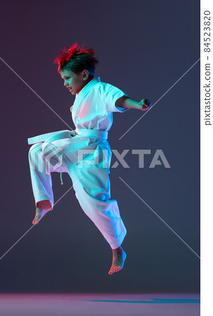 Full-length portrait of boy, karateka in white kimono, jumping, training isolated over gradient blue background in neon lights 84523820