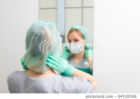young woman doctor in gloves looks in the mirror and adjusts the cap on her head young woman doctor in gloves looks in the mirror and adjusts the cap on her head 84526586