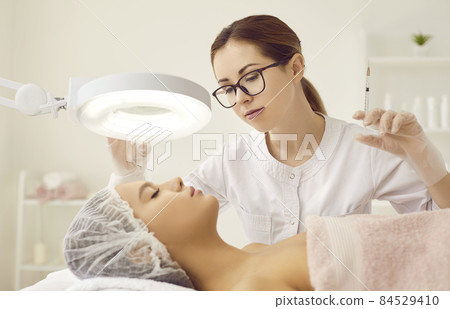 Woman who works as esthetician giving injection or hyaluronic acid filler to client under lamp magnifier. Youthful lady getting face treatment at beauty salon or clinic of aesthetic medicine 84529410