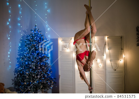Seductive santa in a red bikini dances striptease. Beautiful woman in santa claus hat stands by the pole against the background of the Christmas tree. 84531575