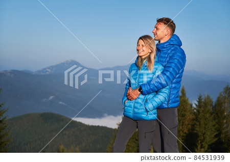 Handsome man hugging his smiling girlfriend from behind and they together standing and watching the surrounding nature against the mountain hills on sunny day. 84531939