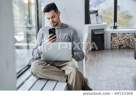 Guy looking at screen of his gadget while sitting at windowsill and working at the laptop 84533369
