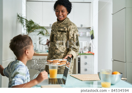 Woman in military uniform offer cookies to her little son while he sitting at the table 84534594