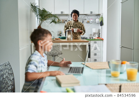 Boy sitting at the table and using laptop while his mother preparing breakfast 84534596